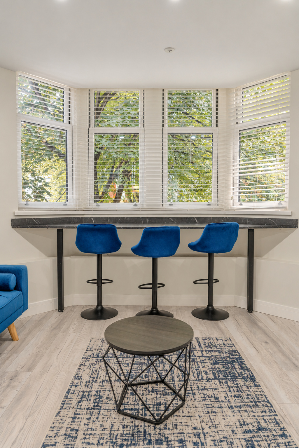Three blue stools beneath a bay-window counter, with a blue sofa and patterned rug in the foreground.