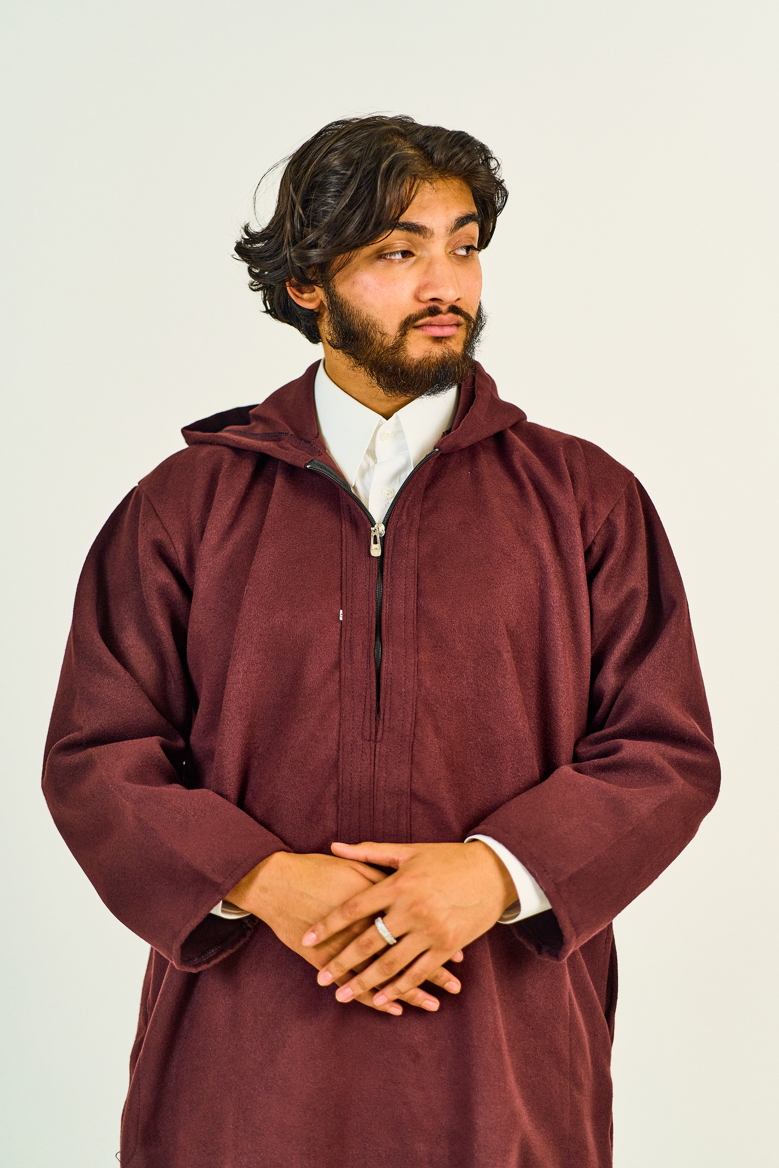A model in a burgundy garment, turned towards the light with his hands resting together.