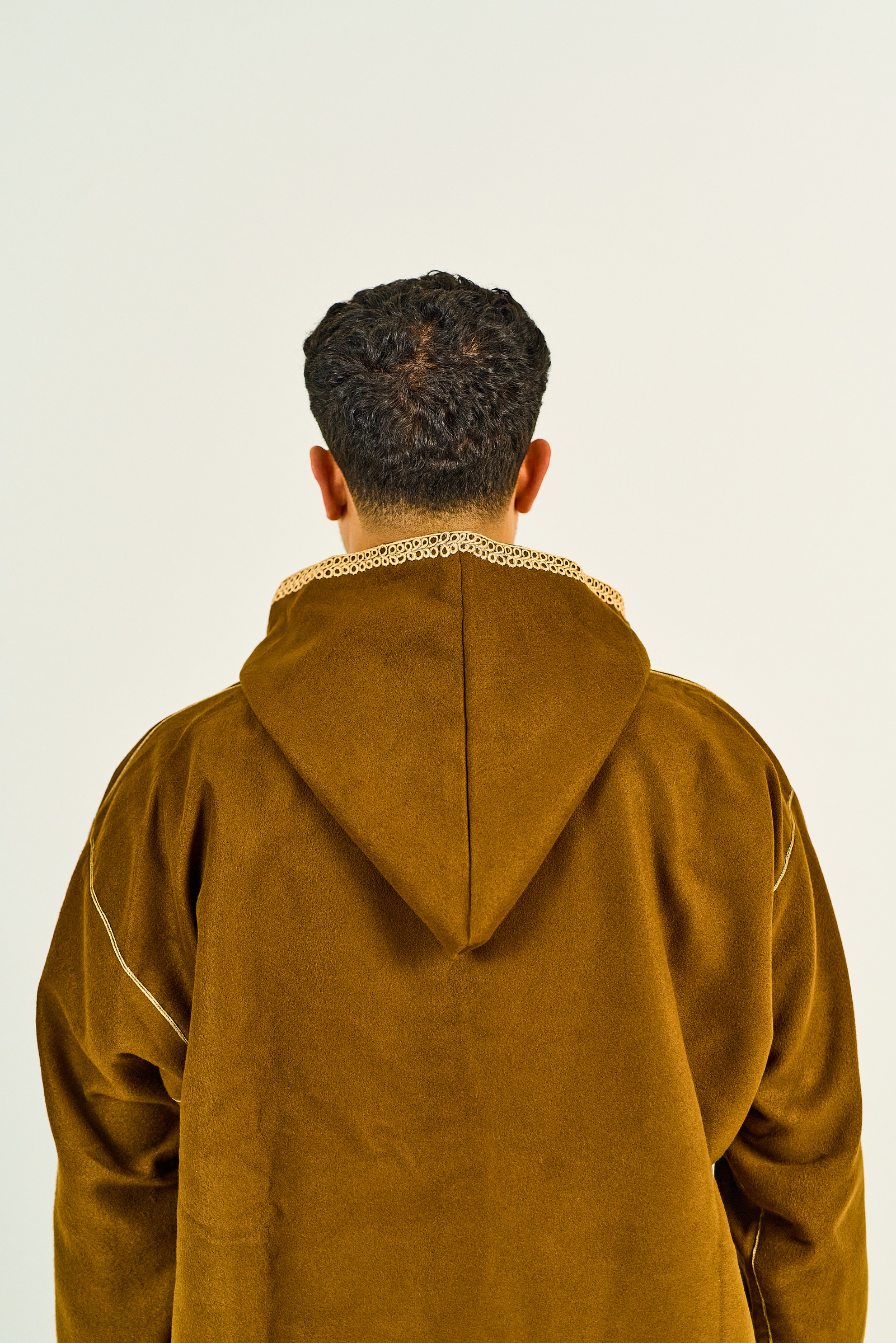 The back of an ochre hooded garment, showing the fabric drape, hood seam and embroidered cream collar.