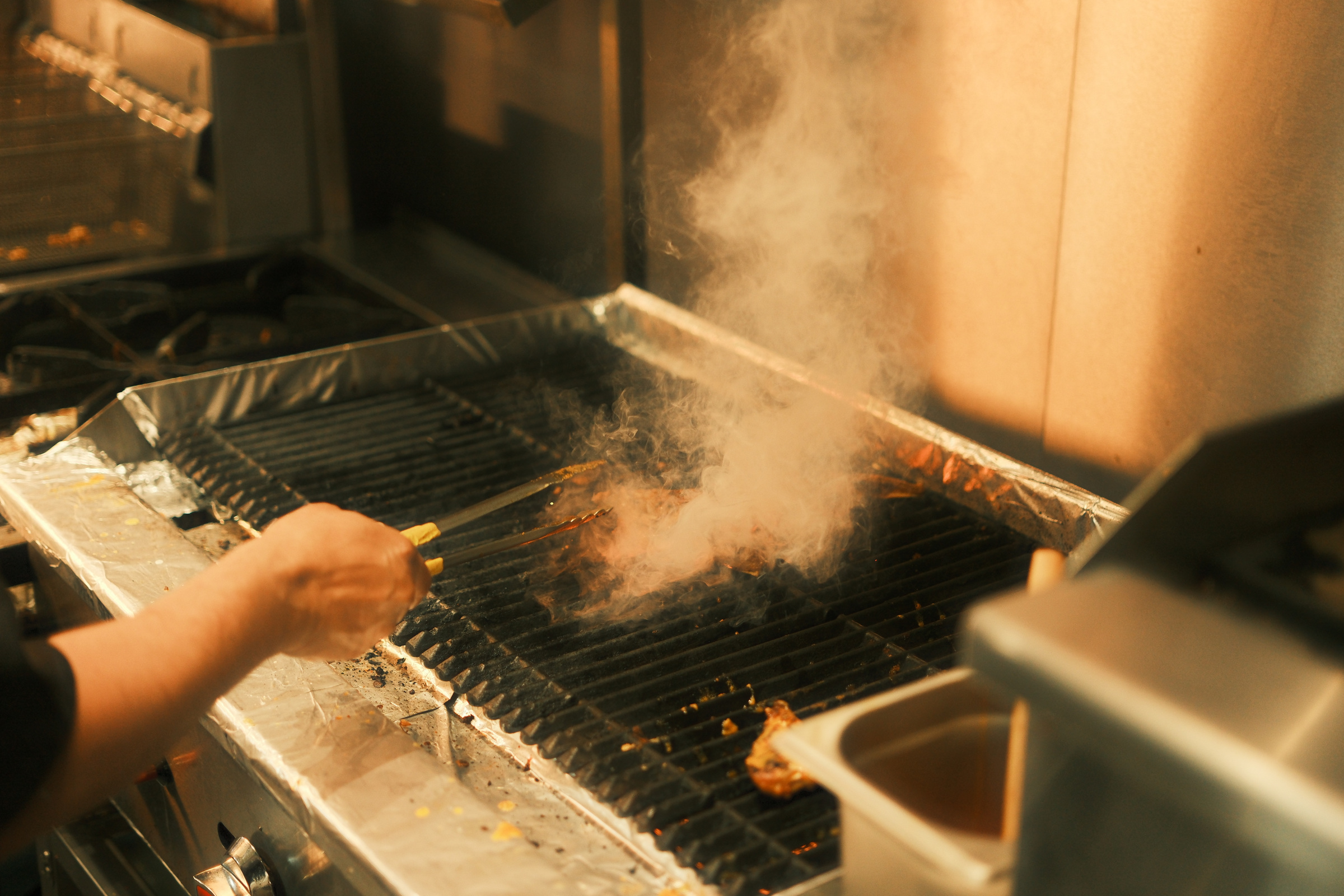 A cook’s hand and tongs over the hot grill, with real kitchen smoke catching warm light.
