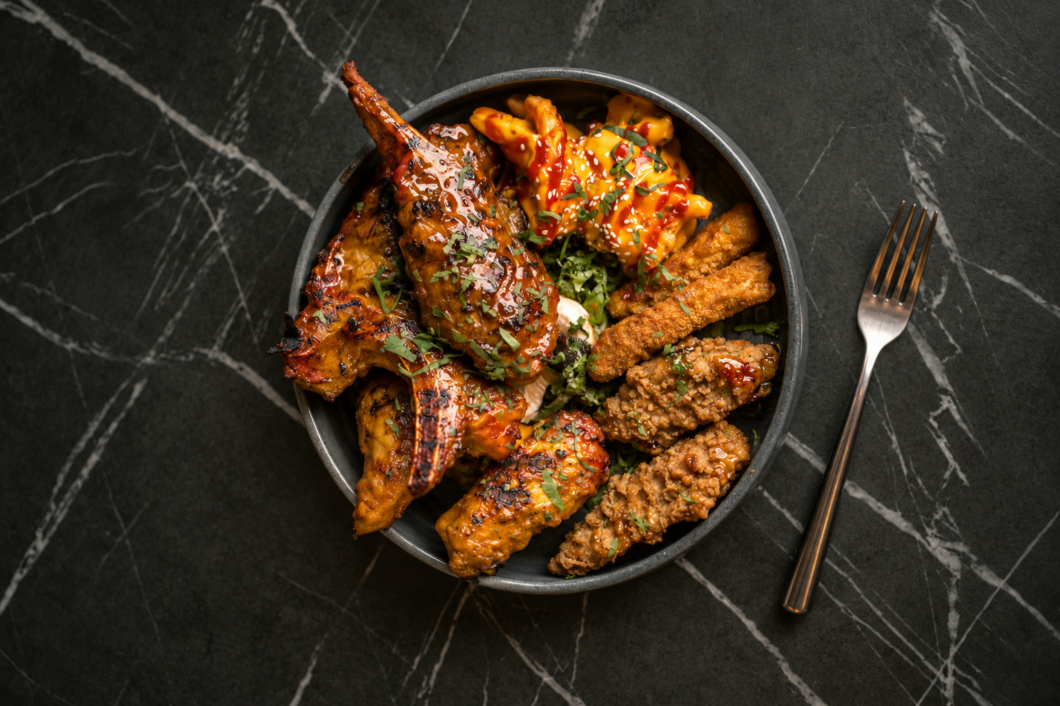 Art-directed overhead chicken sharing bowl with glossy grilled chicken, fries and crisp chicken, on charcoal marble.