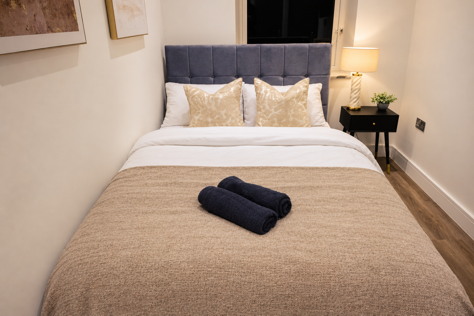 A softly lit guest bed with pale cushions, a textured throw and neatly rolled dark towels.