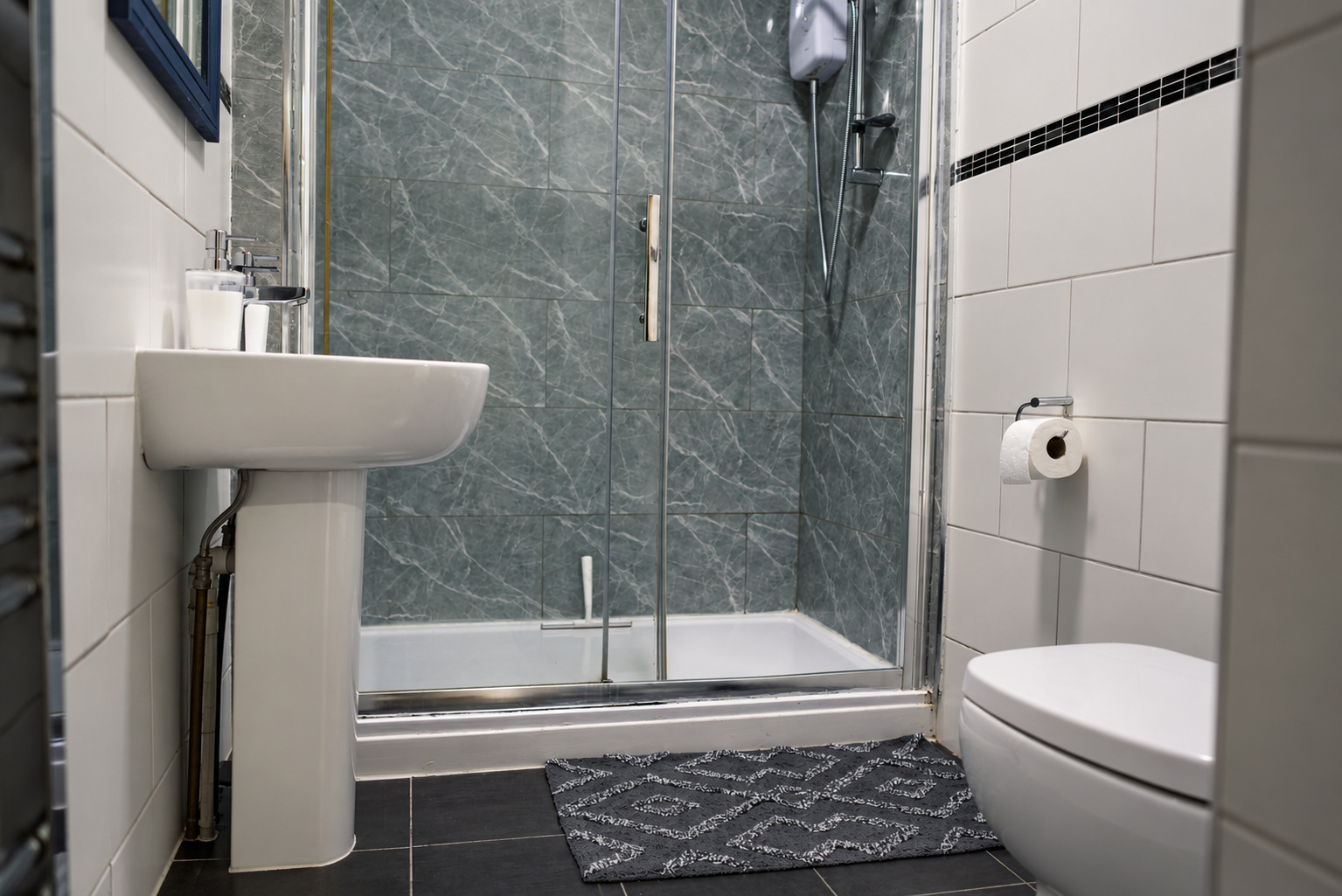A compact shower room with stone tiles, glass enclosure, pedestal basin and patterned bath mat.