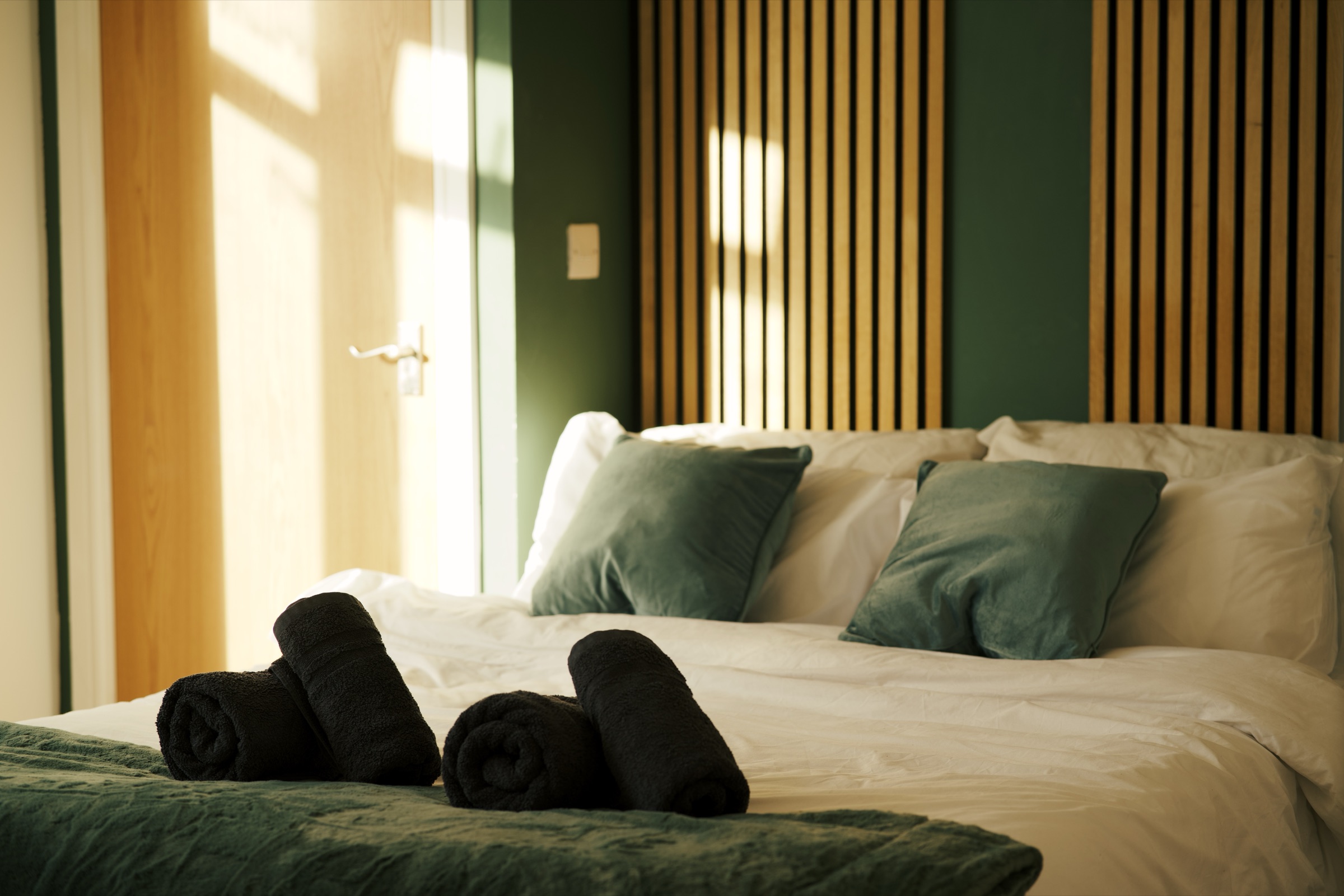 A dressed double bed with rolled towels at its foot, against an oak-slatted headboard wall, in low afternoon light